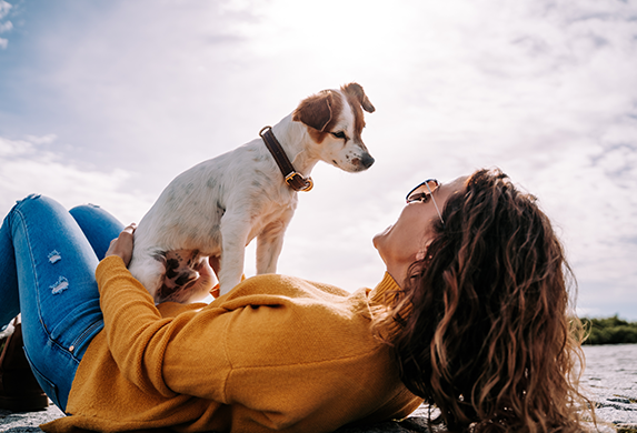 Eine Frau in einem senfgelben Pullover liegt am Strand. Ihr Jack Russel Terrier sitzt auf ihrem Bauch und schaut sie von oben herab an.